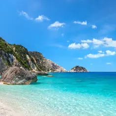 Słoneczna plaża z białym żwirem i turkusową wodą. W oddali skaliste zbocza porośnięte zielenią, charakterystyczne dla greckiej wyspy Kefalonia.