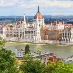 Widok na Parlament w Budapeszcie, cel podróży pociągiem do Budapesztu z Polski. Rzeka Dunaj i zielone drzewa na pierwszym planie.