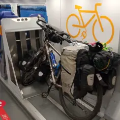 Rowerzysta z bagażami czeka na podróż pociągiem do Włoch. W wagonie rowerowym miejsca dla pasażerów i stojak na rowery.