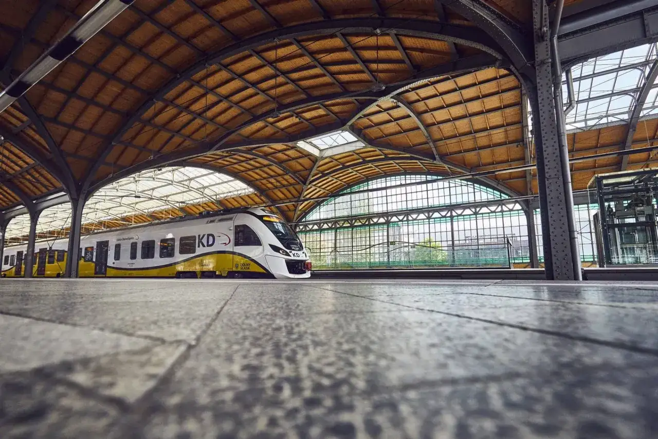 Nowoczesny pociąg KD na peronie dworca PKP Brzeg pod zabytkowym, drewnianym zadaszeniem.