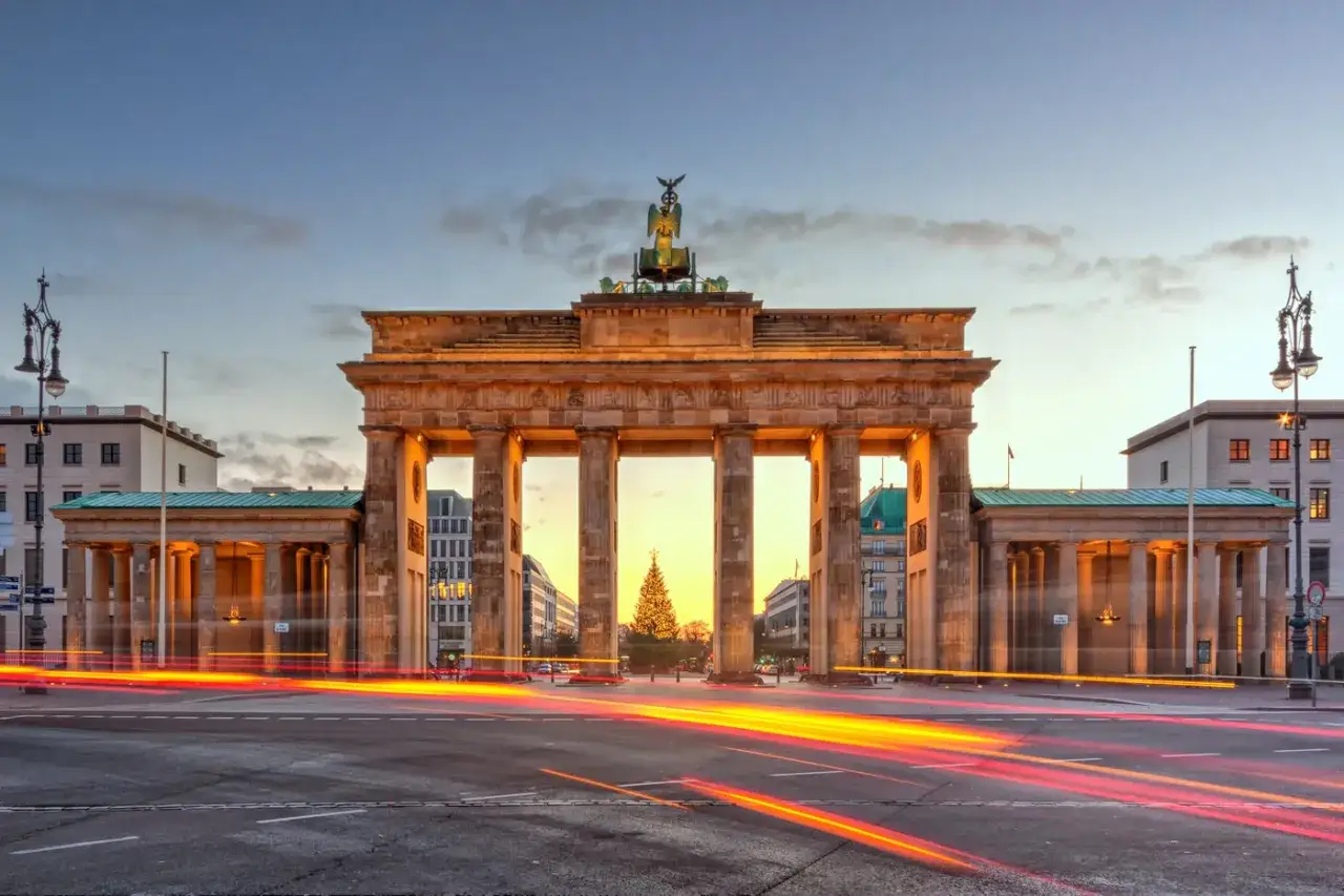 Brama Brandenburska w Berlinie o zmierzchu, z widocznym w tle świątecznym drzewkiem i smugami świateł samochodowych.