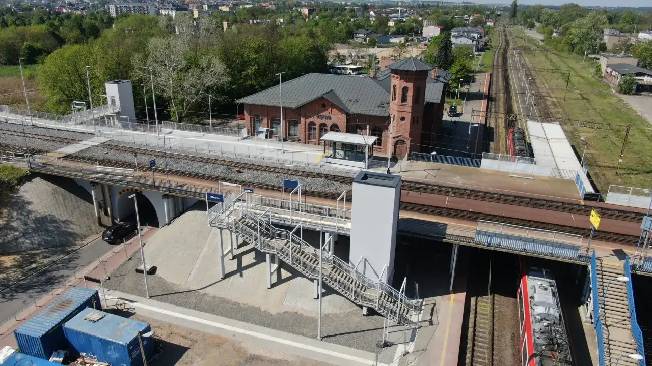 Nowoczesny dworzec kolejowy Kluczbork z ceglanym budynkiem i wieżą. Widoczne tory, perony, kładka i tunel.
