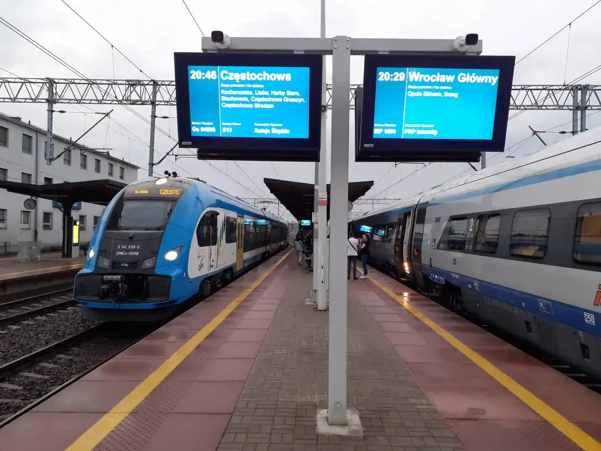 Na dworcu PKP Lubliniec stoją dwa pociągi. Jeden niebieski, drugi szaro-niebieski. Wyświetlacze pokazują odjazdy do Częstochowy i Wrocławia.