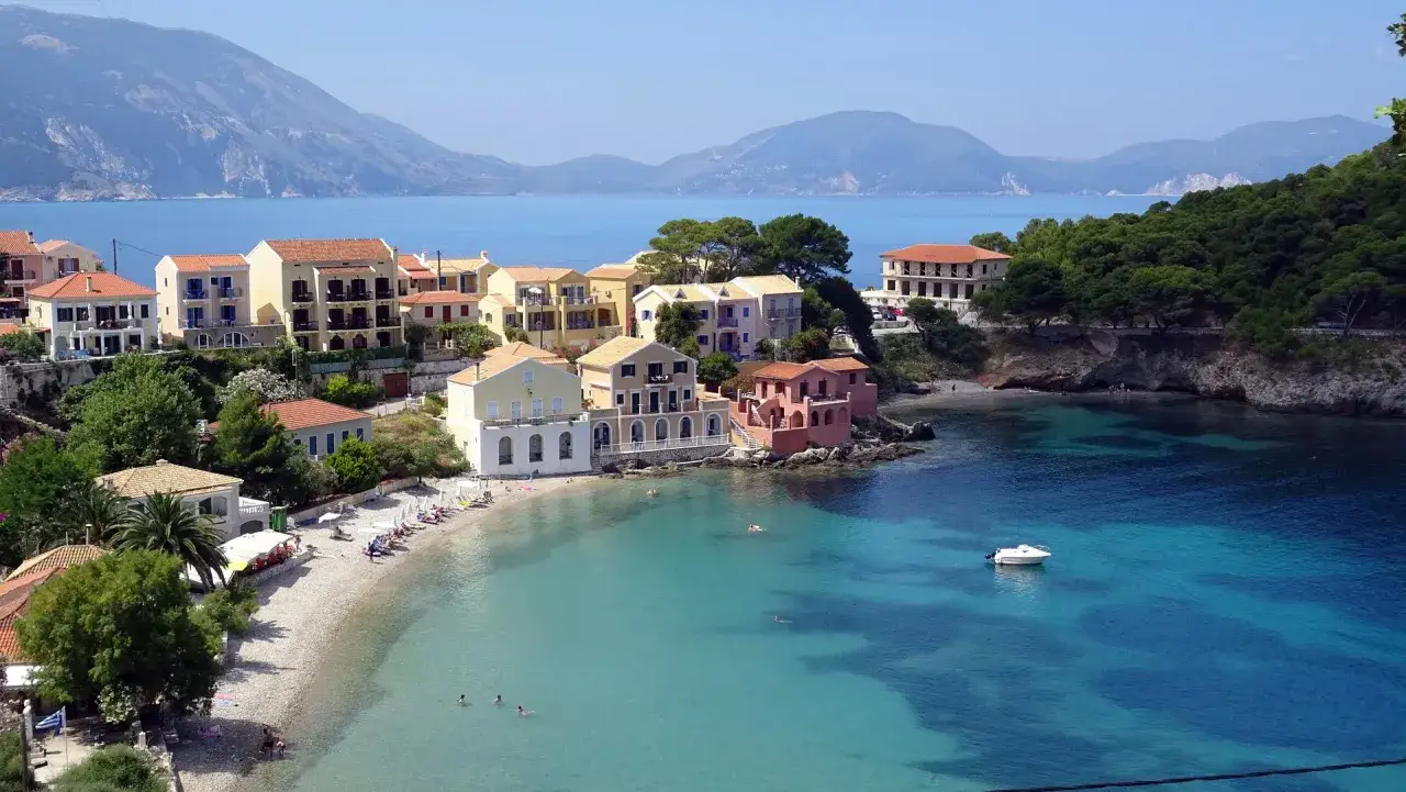 Kolorowe domy nad zatoką na greckiej wyspie Kefalonia. Turkusowa woda, plaża i góry w tle.